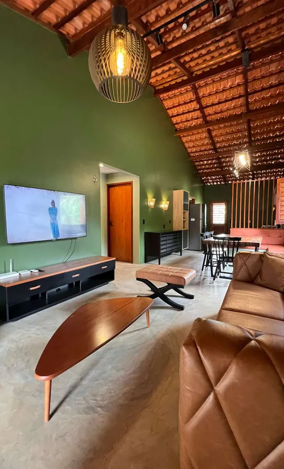 Sala com parede verde, telhado de telha aparente, pendente de arame e tapete claro