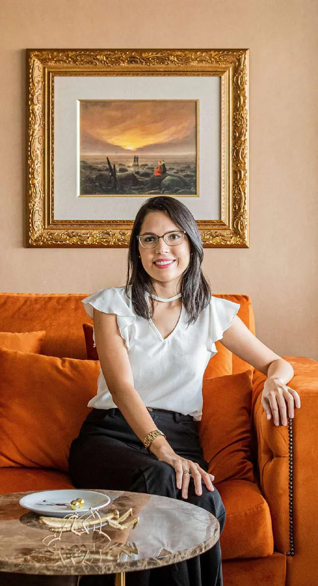 Rhara Assef sentada em um sofá de veludo laranja, sob um quadro de moldura dourada