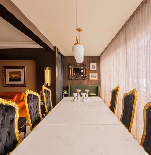 Sala de jantar com cadeiras de encosto dourado, pendente de vidro branco e painel de madeira